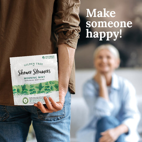 Person holding a Gilded Tree Shower Steamers package with a blurred person in the background and text 'Make someone happy!'.
